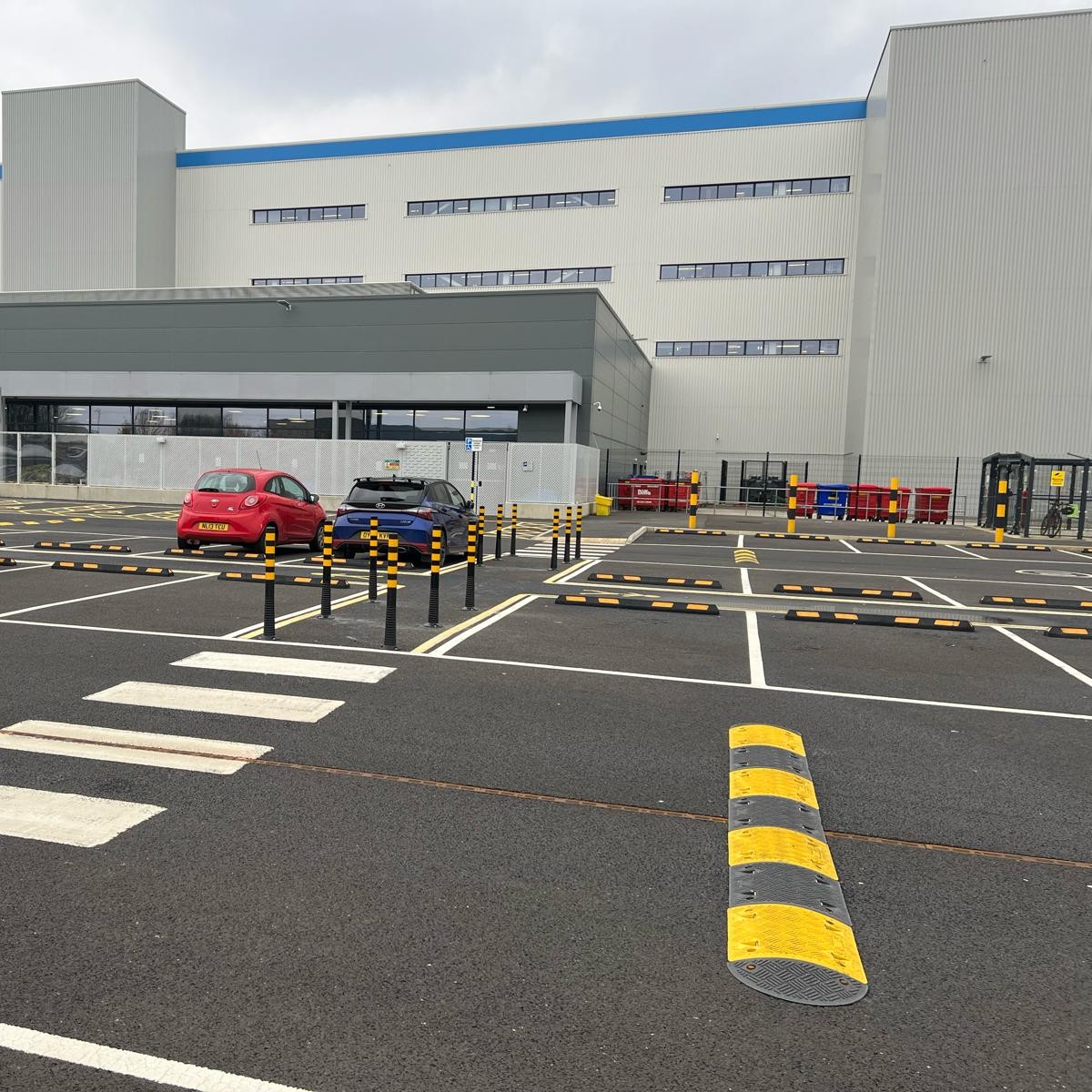 Brandsafe black and yellow flexible bollard delineators installed alongside parking stops and speed bumps in an Amazon vehicle parking and traffic management area.